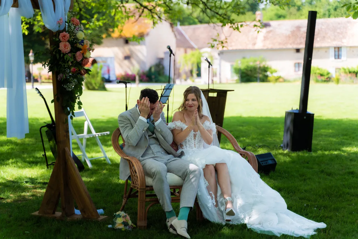 Gerard en Dinah zitten tijdens hun buitenceremonie in rieten stoelen onder de bomen; Gerard slaat zijn handen voor zijn gezicht en Dinah lacht