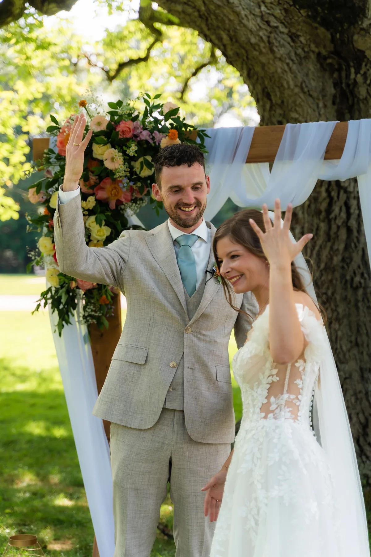 Gerard en Dinah steken lachend hun hand met trouwring in de lucht, onder de ceremonieboog met bloemen
