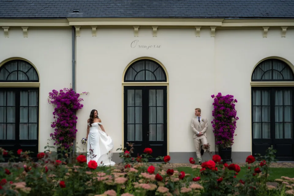 Bruid en bruidegom poseren tegenover elkaar bij de orangerie van Kasteel De Vanenburg, omringd door bloemen
