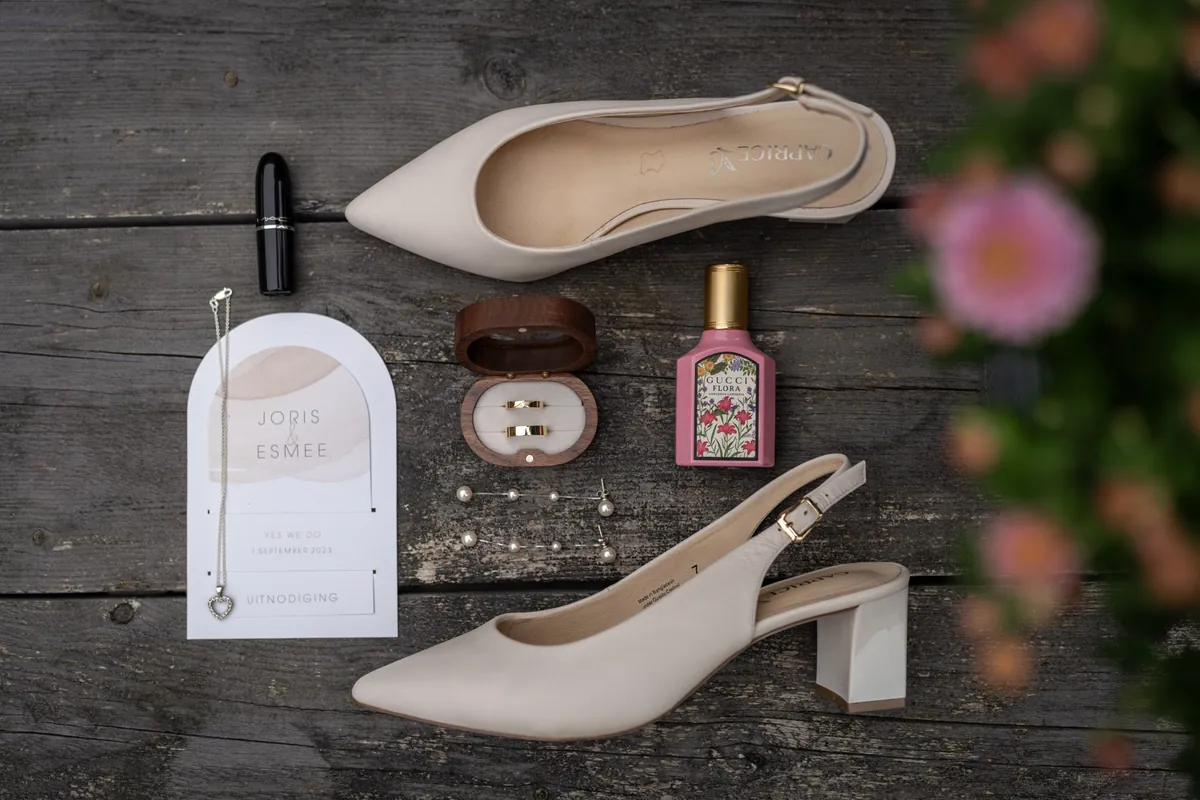 Bruidsaccessoires: schoenen, sieraden, parfum, lipstick en trouwringen op een houten ondergrond.