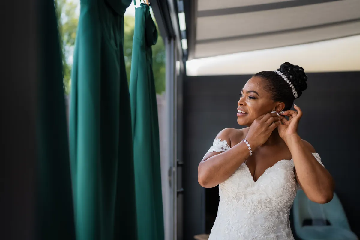 Elegante bruid doet haar oorbellen in bij een raam met groene bruidsmeisjes jurken.