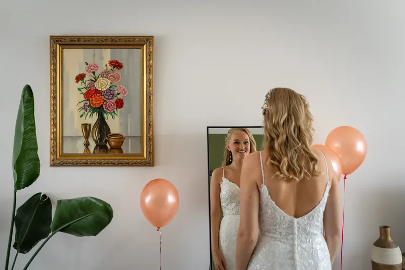 Lachende bruid bekijkt zichzelf in de spiegel met ballonnen en een schilderij op de achtergrond.