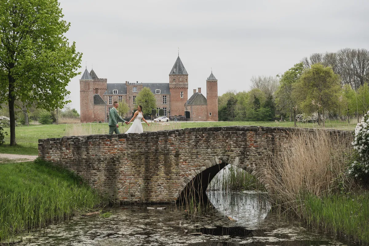 Romantisch bruidspaar loopt hand in hand over een stenen boogbrug met een kasteel op de achtergrond tijdens de Love Shoot.