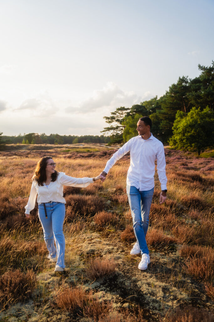 Intieme pre-wedding fotoshoot van een aanstaand bruidspaar dat hand in hand door de heide wandelt tijdens de schemering.