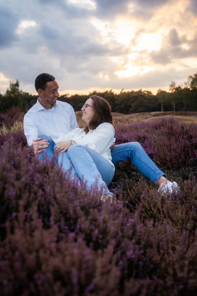 Romantische pre-wedding fotoshoot van een aanstaand bruidspaar zittend in de bloeiende heide tijdens zonsondergang.